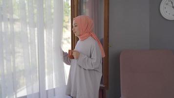 joven mujer en un Pañuelo es acecho fuera el ventana a hogar. joven musulmán mujer mirando fuera ventana a hogar con café jarra en mano. video
