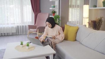 giovane musulmano ragazza studiando a casa. musulmano adolescenziale ragazza Lavorando su il computer portatile a casa e assunzione Appunti nel libro. video