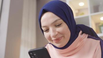 satisfecho musulmán niña vistiendo hijab es utilizando teléfono inteligente a hogar. musulmán niña mirando a su teléfono inteligente video