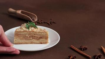 Man's Hand Puts up white plate of Fresh delicious Cake Napoleon with cream on a Brown Background. A cinnamon stick, badyan, coffee beans on a Brown table. Copy space video