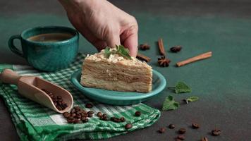 Man's hand Picks up turquoise plate of Fresh delicious Cake Napoleon with cream on a Green Background. A cinnamon stick, badyan, coffee beans on a Green table. Copy space video