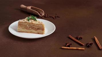 Delicious Napoleon Cake with cream on a white plate decorated with a sprig of mint on a Brown Background. A cinnamon stick, badyan, coffee beans on a brown background. Copy space video