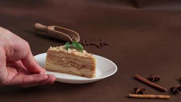 Man's hand Picks up white plate of Fresh delicious Cake Napoleon with cream on a Brown Background. A cinnamon stick, badyan, coffee beans on a Brown table. Copy space video