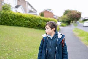 lado ver retrato niño mirando afuera, positivo niño chico caminando a colegio en el mañana, colegio chico en pie solo con borroso casa y la carretera foto