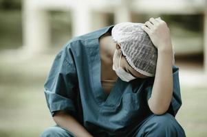 Tired depressed female asian scrub nurse wears face mask blue uniform sits on hospital floor,Young woman doctor stressed from hard work photo