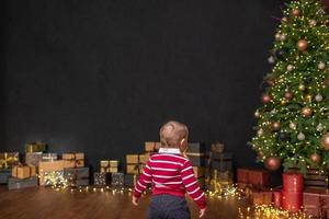 a little boy stands in front of a Christmas tree and boxes with gifts standing along a black wall photo