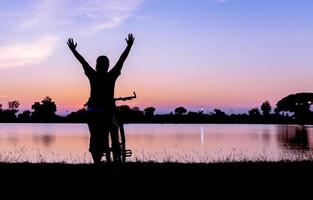 silhouette woman hands up with bicycle on sunset background photo
