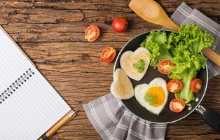 fried eggs and bread in the shape of a heart and fresh vegetables with blank notebook. photo