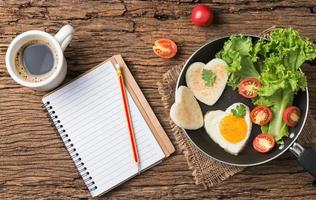 fried eggs and bread in the shape of a heart and fresh vegetables with blank notebook and black coffee photo