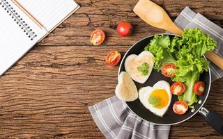 fried eggs and bread in the shape of a heart and fresh vegetables with blank notebook. photo