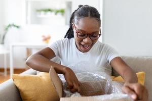 Young satisfied happy african girl woman lady shopaholic customer sit on sofa unpack parcel delivery box, online shopping shipment concept. photo