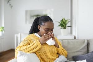 Ill african young woman covered with blanket blowing running nose got fever caught cold sneezing in tissue sit on sofa, sick allergic black girl having allergy symptoms coughing at home, flu concept photo