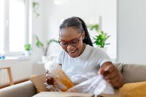 Smiling African woman sit on couch at home open post package shopping online buying goods on internet, happy young female customer unpack postal shipping parcel satisfied with order or delivery photo