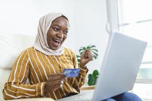 mujer musulmana elegante y atractiva que usa una computadora portátil móvil que busca información de compras en línea en la sala de estar en casa. retrato de mujer feliz comprando productos a través de compras en línea. pagar con tarjeta de crédito