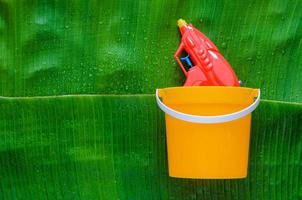 Red water gun in yellow bucket on wet banana leaf background for Thailand Songkran festival. photo