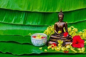 Buddha statue with bowl of water and flowers to do blessing for Thailand Songkran Festival on wet banana leaves background.