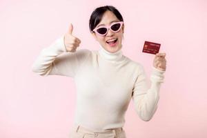 Fascinating fun joyful young woman of Asian ethnicity 20s years old with wear sunglasses wears white shirt hold in hand credit bank card isolated on plain pastel light pink background studio portrait. photo