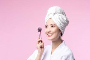 Portrait happy young asian woman with natural make up face holding cosmetic skin powder blusher isolated on pink background. Female apply skincare brush treatment. beauty product, cosmetology concept. photo