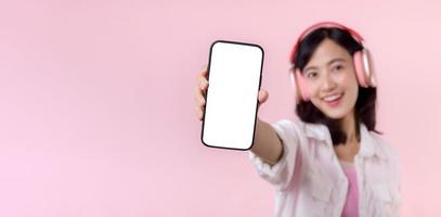 Happy cheerful smiling asian woman with wireless earphones showing blank screen mobile phone or new smartphone music application advertisement mockup isolated on pink studio background.