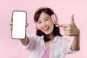 contento alegre sonriente asiático mujer con inalámbrico auriculares demostración blanco pantalla móvil teléfono o nuevo teléfono inteligente música solicitud anuncio Bosquejo aislado en rosado estudio antecedentes. foto