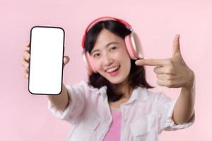 Happy cheerful smiling asian woman with wireless earphones showing blank screen mobile phone or new smartphone music application advertisement mockup isolated on pink studio background. photo