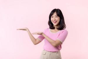 Portrait young beautiful asian woman happy smile pointing finger and hand to showing on copy space isolated on pink background. Attractive female person gesture attention to this promotion. photo