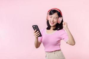 Portrait cheerful young asian woman enjoy listening audio by smartphone music application against pink. Happy smiling female person with headphone. Sound, leisure, lifestyle, technology concept