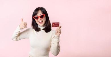 Fascinating fun joyful young woman of Asian ethnicity 20s years old with wear sunglasses wears white shirt hold in hand credit bank card isolated on plain pastel light pink background studio portrait. photo