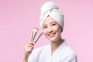 Portrait happy young asian woman with natural make up face holding cosmetic skin powder blusher isolated on pink background. Female apply skincare brush treatment. beauty product, cosmetology concept. photo