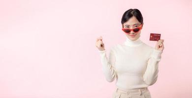 Fascinating fun joyful young woman of Asian ethnicity 20s years old with wear sunglasses wears white shirt hold in hand credit bank card isolated on plain pastel light pink background studio portrait. photo