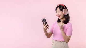 retrato alegre joven asiático mujer disfrutar escuchando audio por teléfono inteligente música solicitud en contra rosado. contento sonriente hembra persona con auricular. sonido, ocio, estilo de vida, tecnología concepto foto