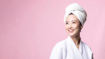 hermosa joven asiático mujer contento con limpiar cara piel en toalla y bata de baño, spa traje en rosado antecedentes. protección de la piel, tratamiento, bienestar terapia, facial cuidado, belleza hembra salud, cosmetología concepto foto