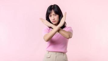 portrait serious young asian woman with cross arm gesture showing stop, no, wrong, denial, rejection sign isolated on pink pastel studio background. deny and negative expression symbol concept. photo