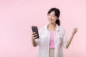 Pretty young asian woman showing success, victory hand gesture while receiving great news from smartphone on pink background. Happy technology, mobile phone advertisement, online application concept photo