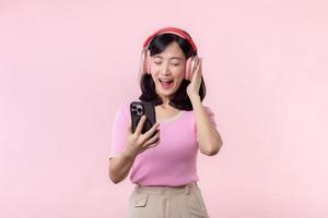 Portrait cheerful young asian woman enjoy listening audio by smartphone music application against pink. Happy smiling female person with headphone. Sound, leisure, lifestyle, technology concept photo