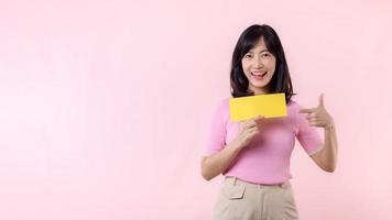 Portrait happy young woman model holding and showing blank space paper for advertisement information message poster with thumb up or point finger gesture isolated on pink pastel studio background. photo