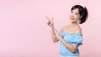 Portrait young beautiful asian woman happy smile pointing finger and hand to showing on copy space isolated on pink background. Attractive female person gesture attention to this promotion. photo