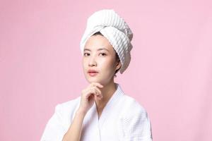 hermosa joven asiático mujer contento con limpiar cara piel en toalla y bata de baño, spa traje en rosado antecedentes. protección de la piel, tratamiento, bienestar terapia, facial cuidado, belleza hembra salud, cosmetología concepto foto