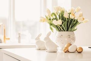 Pascua de Resurrección mesa ajuste con tulipanes, Pascua de Resurrección conejitos, y huevos con dorado patrones en el blanco estilo escandinavo cocina antecedentes. hermosa minimalista diseño para saludo tarjeta foto