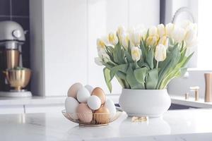 Easter table setting with tulips, Easter bunnies, and eggs with golden patterns in the white Scandinavian-style kitchen background. Beautiful minimalist design for greeting card photo