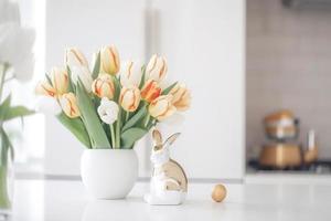 Easter table setting with tulips, Easter bunnies, and eggs with golden patterns in the white Scandinavian-style kitchen background. Beautiful minimalist design for greeting card photo