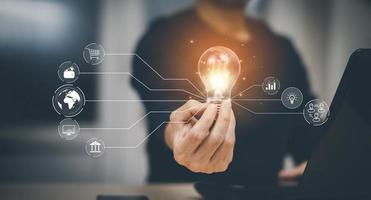 Businessman hand holding light bulb with icons and working on the desk, Creativity and innovation are keys to success.Concept of new idea and innovation with energy and power , working at home, photo