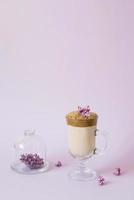 trendy drink 2020 dalgon Coffee in a transparent Cup with a paper tube, a plate with lilac flowers on a lilac background with a copy space photo