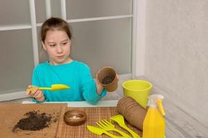 girl pours soil into a peat pot, next to garden tools, a sprinkler, pumpkin seeds, planting seedlings photo