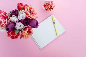 Top view of pink desk with notepad and pen. Bouquet with tulips of different varieties photo