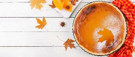 American homemade pumpkin pie with cinnamon and nutmeg, pumpkin seeds and autumn leaves on a white wooden table. Thanksgiving food. Top view. Banner photo