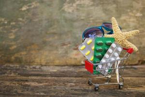 Shopping cart with pills, sunglasses, seashells on old wooden background. Concept of medication required in journey. photo