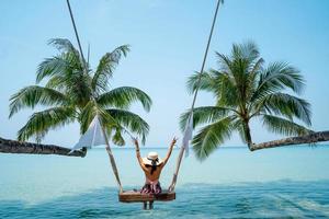 Woman enjoying the nature of the sea beach. She was sitting on a wooden swing under the shade of a palm tree. Travel concept.