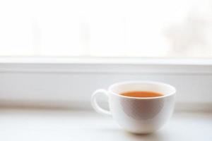 blanco taza con té soportes en el antepecho en el ventana foto