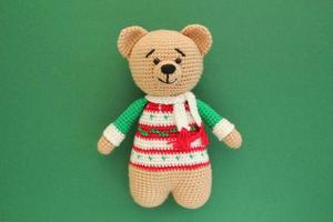 Crochet amigurumi handmade stuffed soft teddy bear toy in colored sweater on green background. Handwork, hobby. Craft diy newborn pregnancy concept. Knitted doll for little baby. Closeup flat lay out photo